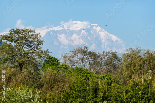 A view on snow capped Himalayas, seen from Phewa Lake in Pokhara, Nepal. Sharp and dangerous slopes. There is a hill in front of it, covered with forest. There is a dence forest at the shore. Serenity