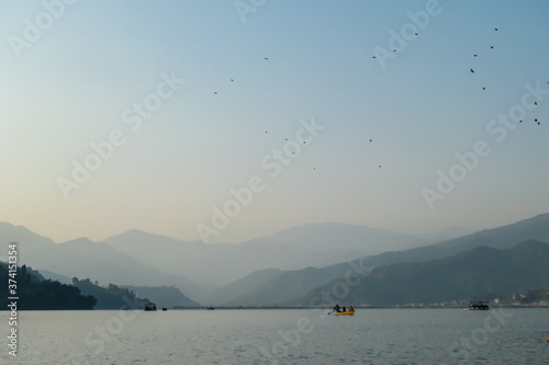 An idyllic view on Phewa Lake in Pokhara, Nepal. There are high Himalayan ranges around the lake. Calm surface of the lake. Clear and sunny day. There is a dence forest at the shore. Golden hour haze