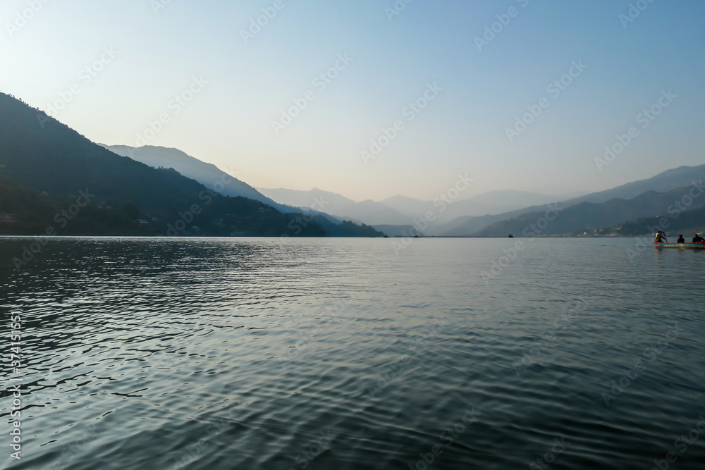 An idyllic view on Phewa Lake in Pokhara, Nepal. There are high Himalayan ranges around the lake. Calm surface of the lake. Clear and sunny day. There is a dence forest at the shore. Golden hour haze