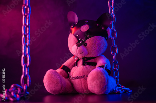 toy bear dressed in leather belts harness accessory for BDSM games on a dark background in neon light