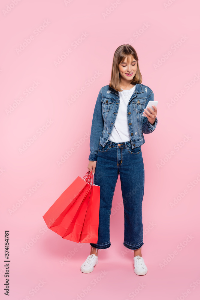 Naklejka premium Woman in jeans and denim jacket using smartphone and holding red shopping bags on pink background
