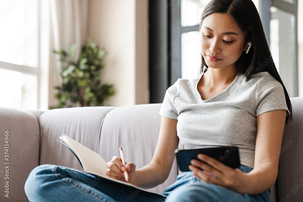 Image of young asian woman using mobile phone while writing down notes Image of young asian woman using mobile phone while writing down notes