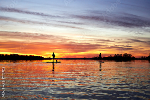 Silhouettes of two people who are paddling on a SUP boards on the autumn Danube river at sunset. Stand up paddle boarding - awesome active recreation in nature. Rear view. Backlight.