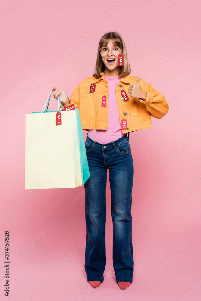 Fototapeta premium Excited woman holding shopping bags with price tags and showing thumb up on pink background