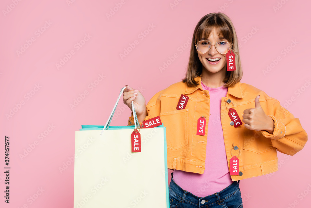 Fototapeta premium Woman with price tags on jacket holding shopping bags and showing like gesture isolated on pink