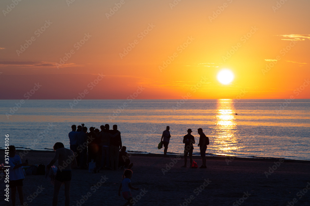 Beautiful sunset at the sea. Young people walk along the beach. People are watching the sunset. Sea and red-yellow sky. Silhouettes of people on the background of the sunset. In the evening by the sea