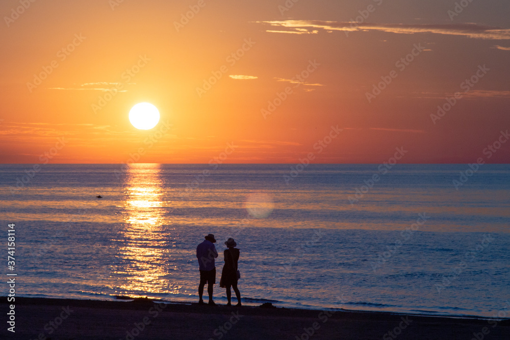 Beautiful sunset at the sea. Young people on the beach. Couple are watching the sunset. Sea and red-yellow sky. Silhouettes of people on the background of the sunset. In the evening by the sea