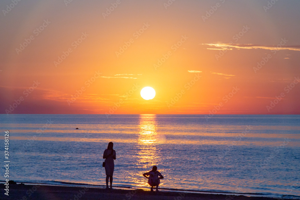 Fototapeta premium Beautiful sunset at the sea. Young people on the beach. Couple are watching the sunset. Sea and red-yellow sky. Silhouettes of people on the background of the sunset. In the evening by the sea