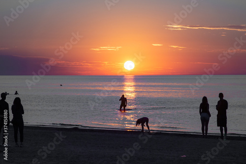 Beautiful sunset at the sea. Young people on the beach. Couple are watching the sunset. Sea and red-yellow sky. Silhouettes of people on the background of the sunset. In the evening by the sea