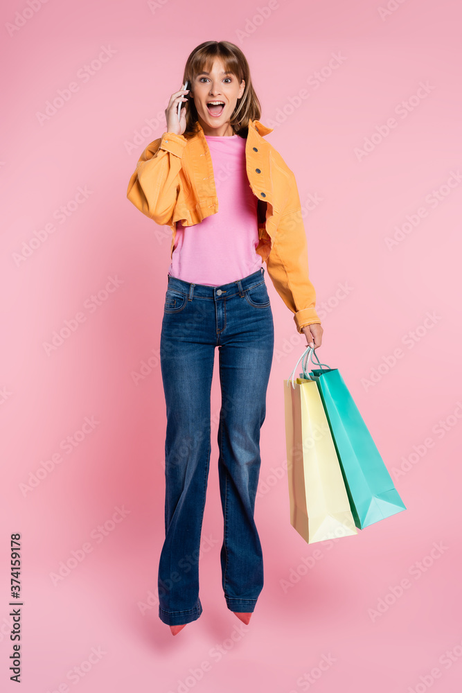 Excited woman holding shopping bags and talking on smartphone while jumping on pink background Excited woman holding shopping bags and talking on smartphone while jumping on pink background