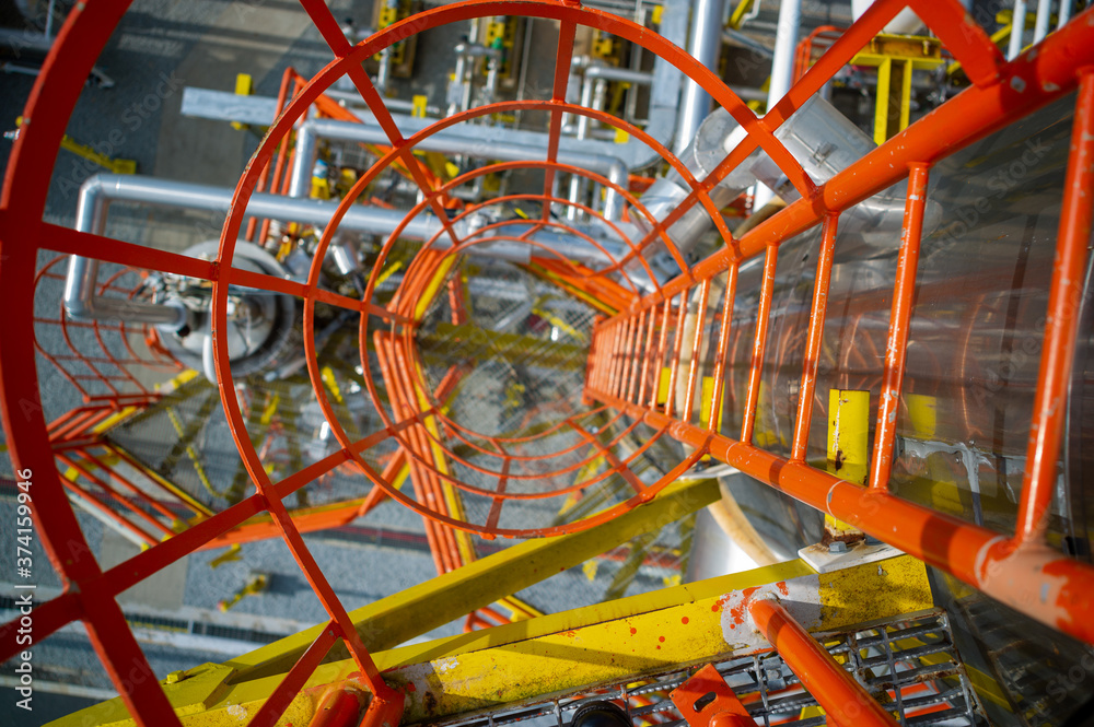 Looking down an industrial steel ladder with safety cage. Heavy industry gas and petroleum plant. Pov with sleective focus and vivid yellow, orange and reflective steel colors as safety first concept.