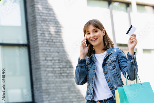 Young woman showing credit card while holding shopping bags and talking on smartphone on urban street