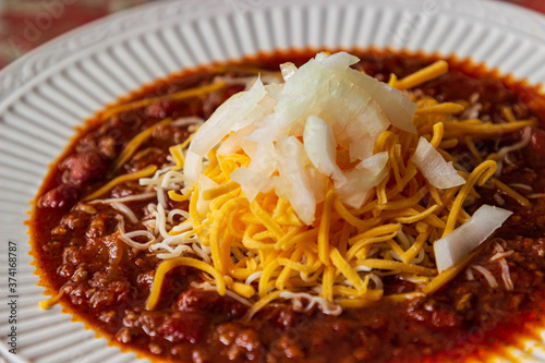 A bowl of delicious homemade chili in a white bowl and garnished with cheddar and Monterrey Jack cheeses and diced onions.