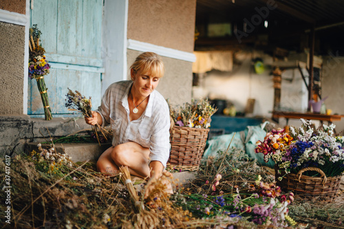 Positive woman in a shirt at the cottage creates bouquets of dried flowers with a smile on his face. Amateur florist makes compositions from flowers in the country.
