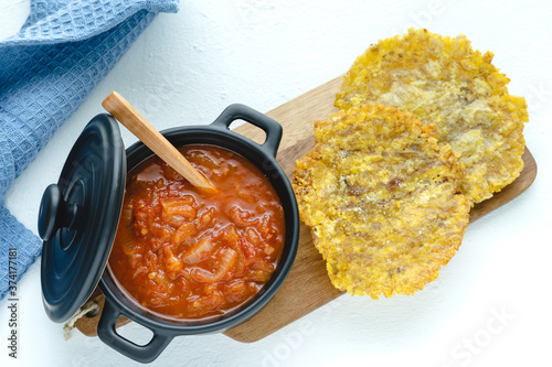 Fototapeta Naklejka Na Ścianę i Meble -  Pot with fried tomato accompanied by fried banana on a white background. Typical dish of Colombian cuisine. Copy space. Patacón con hogao.