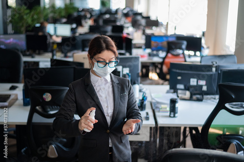 Female manager in a medical mask sprays on hand antiseptic. A woman in a suit uses a sanitizer to disinfect. Concept of office work during the coronavirus epidemic.