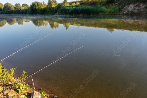 Two fishing rods standing on the sandy shore of a fish pond, lit by the morning sun.