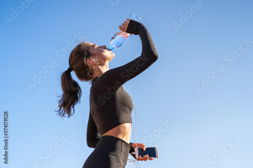 Fototapeta Naklejka Na Ścianę i Meble -  Thirsty young fit woman drinking water from plastic bottle, resting after jogging, holding mobile smartphone in her hand, listens to music. Blue sky with copy space. Hot day. 