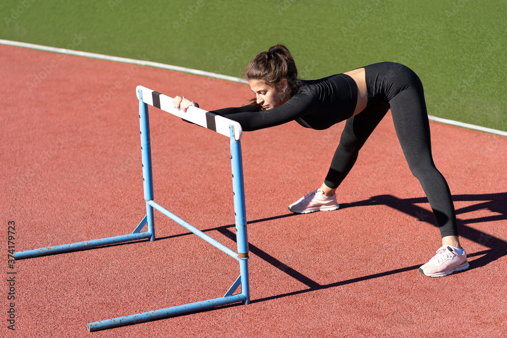 Fit young woman in black sportswear doing strength exercises before running on a treadmill rubber stadium on sunny summer day 