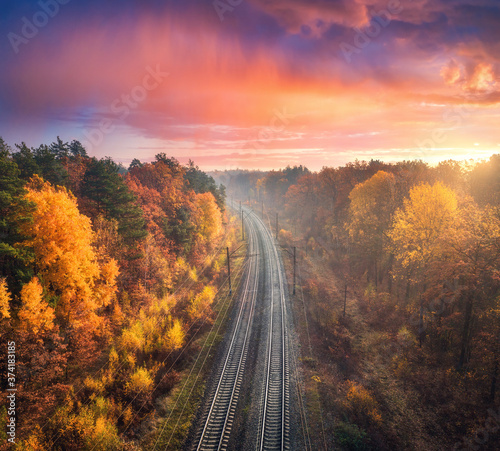 Aerial view of beautiful railroad in autumn forest in foggy sunrise. Industrial landscape with railway station, blue sky with red clouds, trees with orange leaves, fog. Top view of railroad in fall