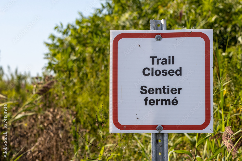 Trail closed sign in English and French with overgrown trail in background