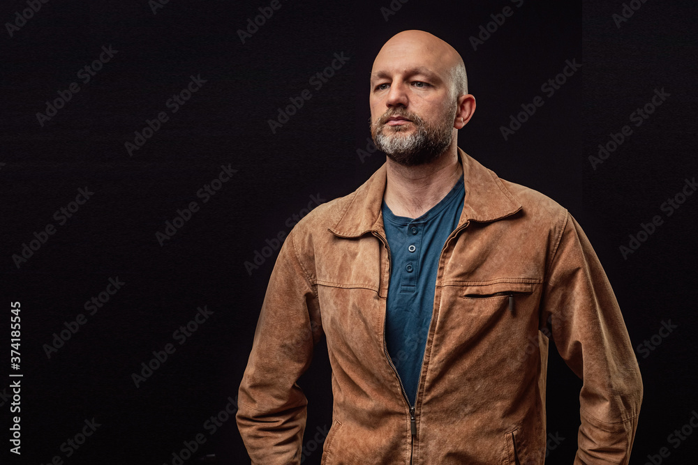 Obraz premium Portrait of a bald man in his 40s dressed in brown suede jacket. Model with grey and beard. Dark background.