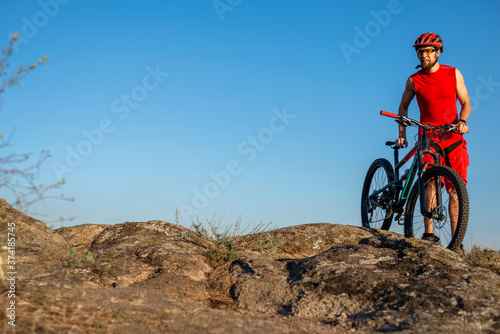 Fototapeta Naklejka Na Ścianę i Meble -  A cyclist in a red jersey and shorts stands on the rock with a bicycle against the blue sky, copy space.