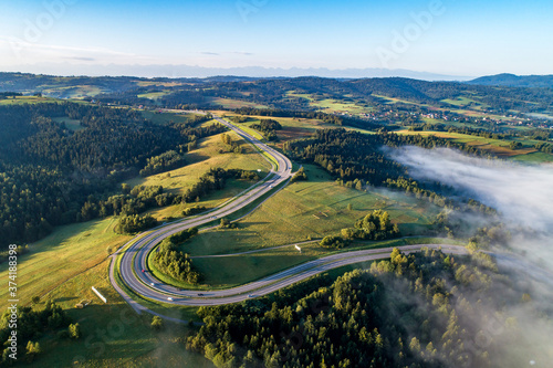 Fototapeta Naklejka Na Ścianę i Meble -  Poland. Winding switchback road from Krakow to Zakopane, called Zakopianka, near Rabka and Chabowka. Aerial view in sunrise light with morning fog. Far view of Tatra mountains in the background