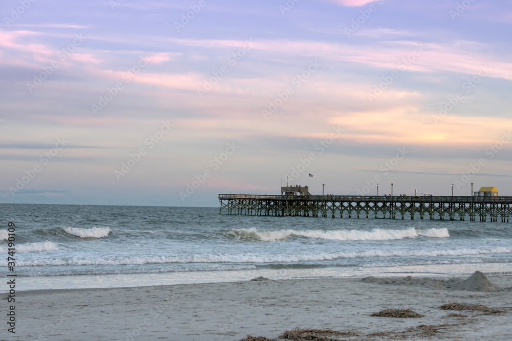 The Apache Pier at sunset in Myrtle Beach South Carolina. Waves are at the foreground with blues and pinks in the sky.
