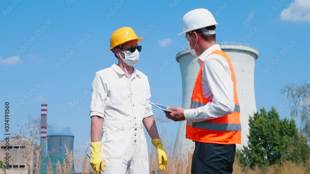 Two masked engineers talk while working on a site. Scheduled maintenance of buildings and structures using a tablet. Two masked engineers talk while working on a site. Scheduled maintenance of buildings and structures using a tablet.
