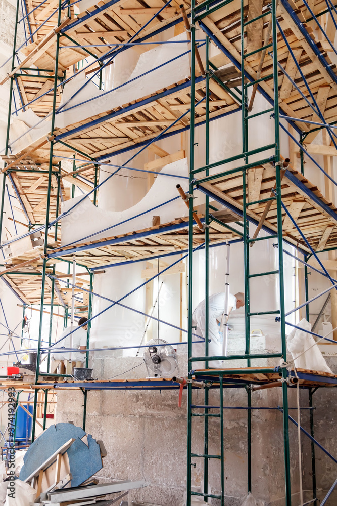 Workers restore white walls on scaffolding in large room. Construction works, interior repairs, restoration and reconstruction of antique buildings are underway