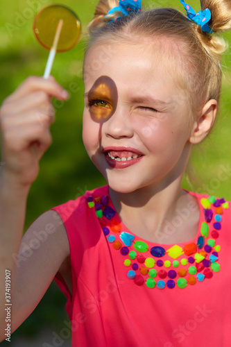 Vertical photo close-up portrait of a cheerful girl in the park. A six-year-old girl with a toothless smile looks at the sun through a transparent lollipop. The girl is wearing a bright pink dress wit
