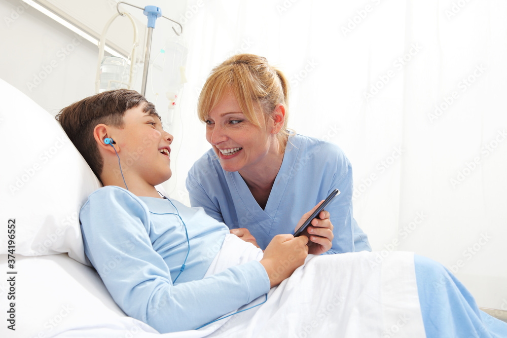 Obraz premium Happy child lying in bed in hospital room and smiling nurse using smartphone surfs the internet wearing earphones