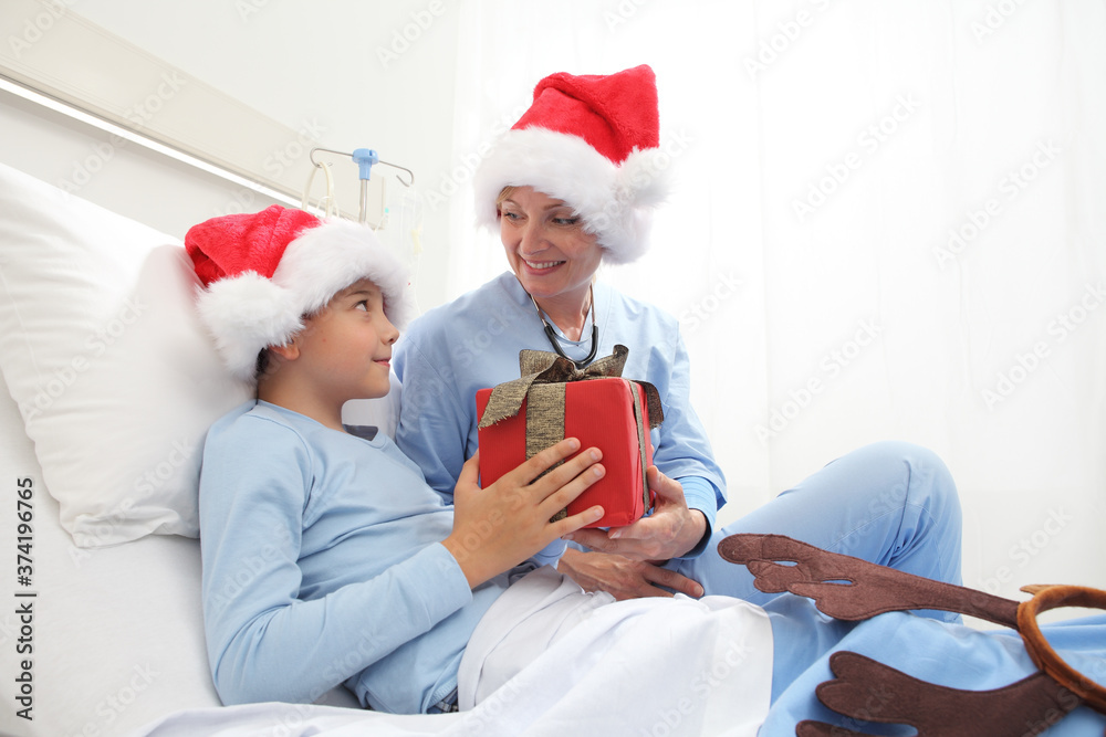 Fototapeta premium Christmas holiday in hospital happy child lying in bed with Santa Claus hat and nurse giving him a present