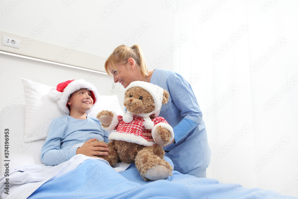 Christmas holiday in hospital happy child lying in bed with Santa Claus hat and nurse dressing a teddy bear Christmas holiday in hospital happy child lying in bed with Santa Claus hat and nurse dressing a teddy bear