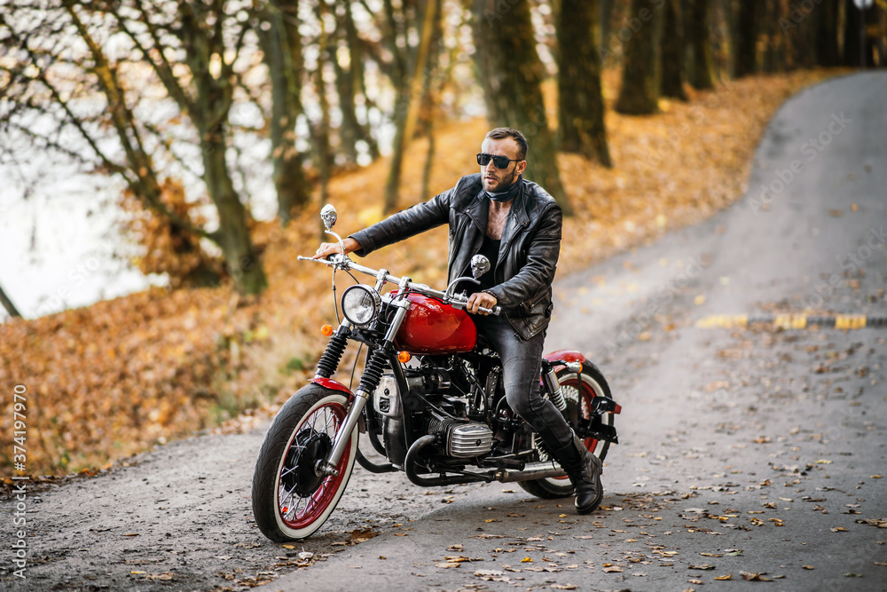 Obraz premium Brodaty brutalny mężczyzna w okularach przeciwsłonecznych i skórzanej kurtce siedzi na motocyklu na drodze w lesie