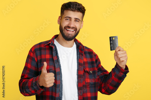 Cheerful bearded hipster young man in plaid shirt holding credit card over yellow background. Man show thumb up.