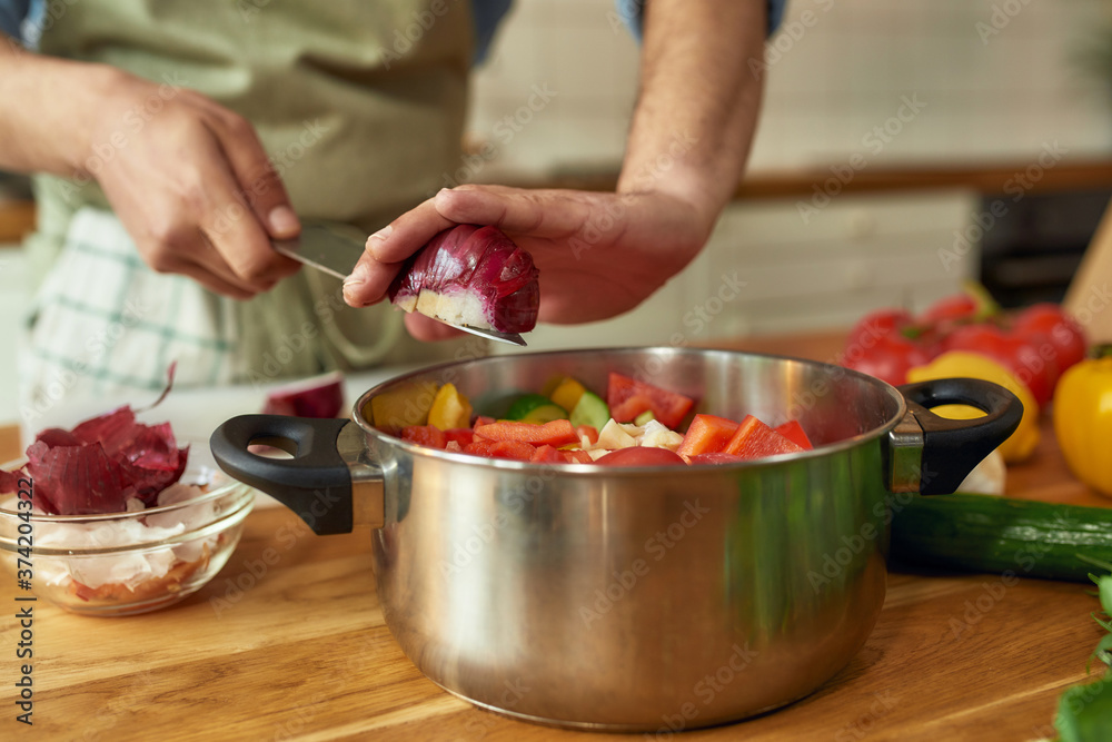Close up of hands of man, chef cook adding onion to the pot with chopped vegetables while preparing a meal in the kitchen