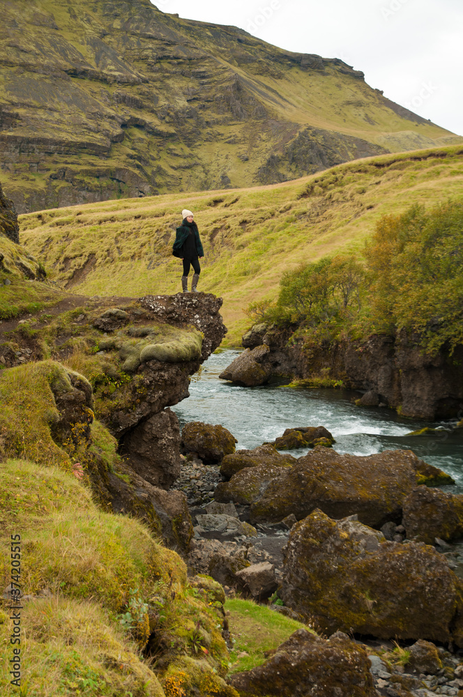 Girl wearing a hat and wellington boots stands on the edge of a rock on top of a river while contemplating the stunning Icelandic landscape