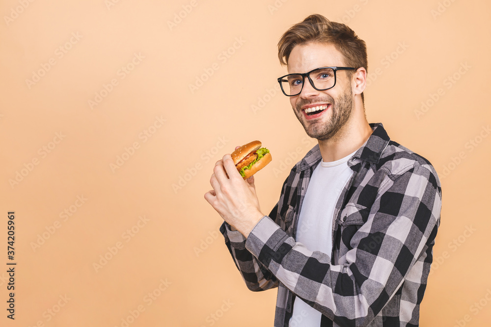 Young man holding a piece of hamburger. Student eats fast food. Burger is not helpful food. Very hungry guy. Diet concept. Isolated over beige background.