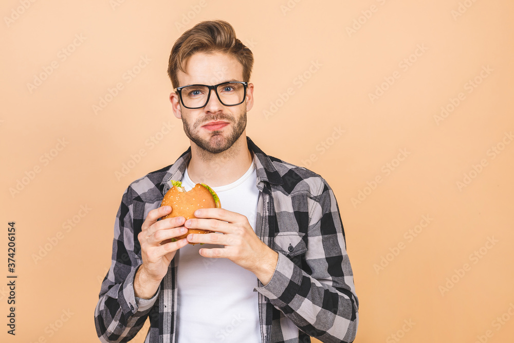 Young man holding a piece of hamburger. Student eats fast food. Burger is not helpful food. Very hungry guy. Diet concept. Isolated over beige background. Young man holding a piece of hamburger. Student eats fast food. Burger is not helpful food. Very hungry guy. Diet concept. Isolated over beige background.