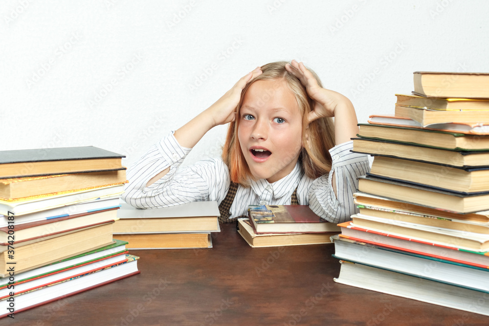 Portrait of a teenage girl sitting at a table tired among the books and doing her homework. Holds his head with his hands. Isolated background. Portrait of a teenage girl sitting at a table tired among the books and doing her homework. Holds his head with his hands. Isolated background.