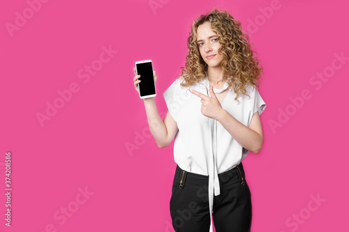 Look at this cell phone. Contented happy woman, pointing her index finger at a blank screen, shows a modern device. Isolated on a pink background.