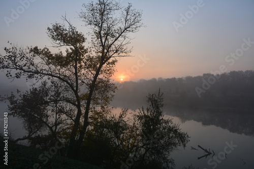 There is a sunrise over the calm and sleeping river on a foggy day in October. 