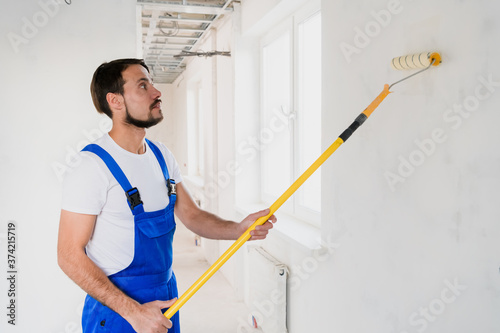 A bearded workman in a blue overalls is painting a wall in the hallway. He uses a roller