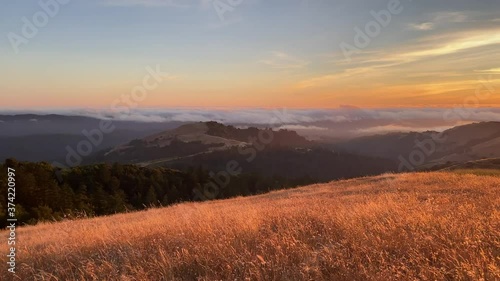 Sunset view of the Santa Cruz mountains, with sun rays illuminating hills covered in dry grass; a layer of clouds visible in the background, covering the coastline; San Francisco Bay Area, California