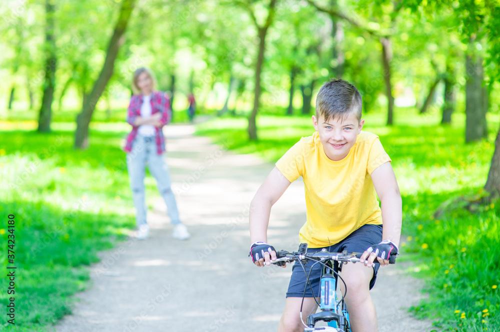Smiling young boy rides on a bicycle in summer sunny park. Mom stands behind on the background