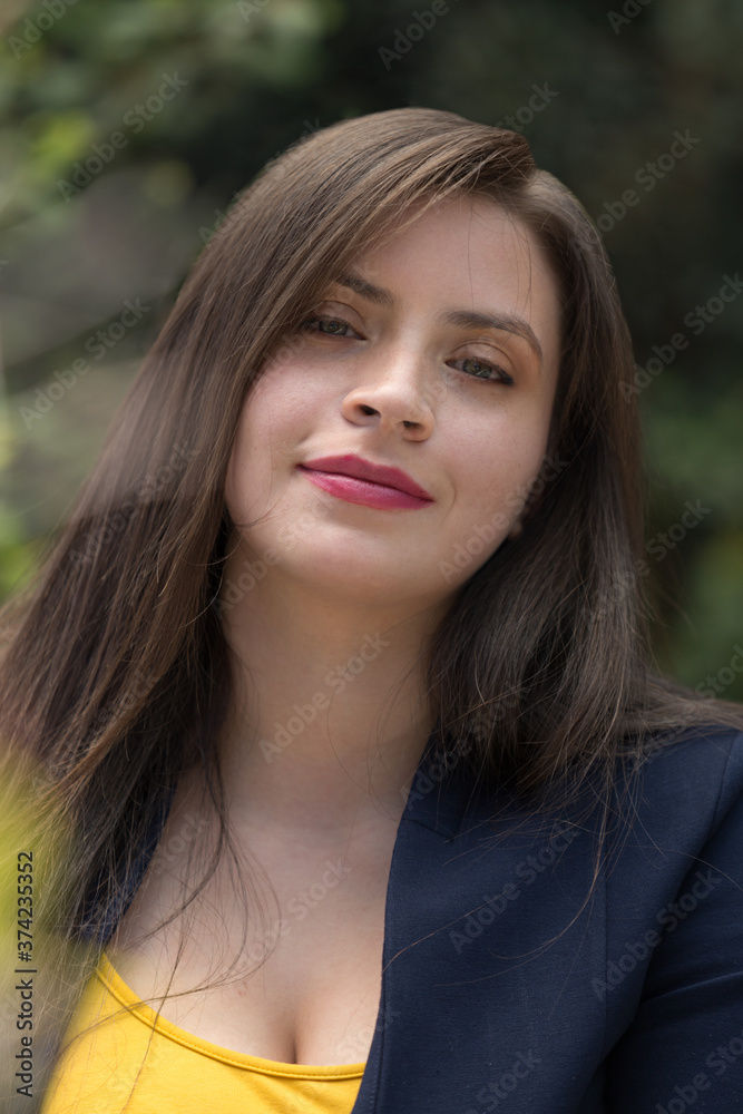 face of young white-skinned Latin woman with long, straight hair, light eyes and makeup, smiling, looking straight ahead, wearing a yellow blouse and blue jacket, photo in natural daylight