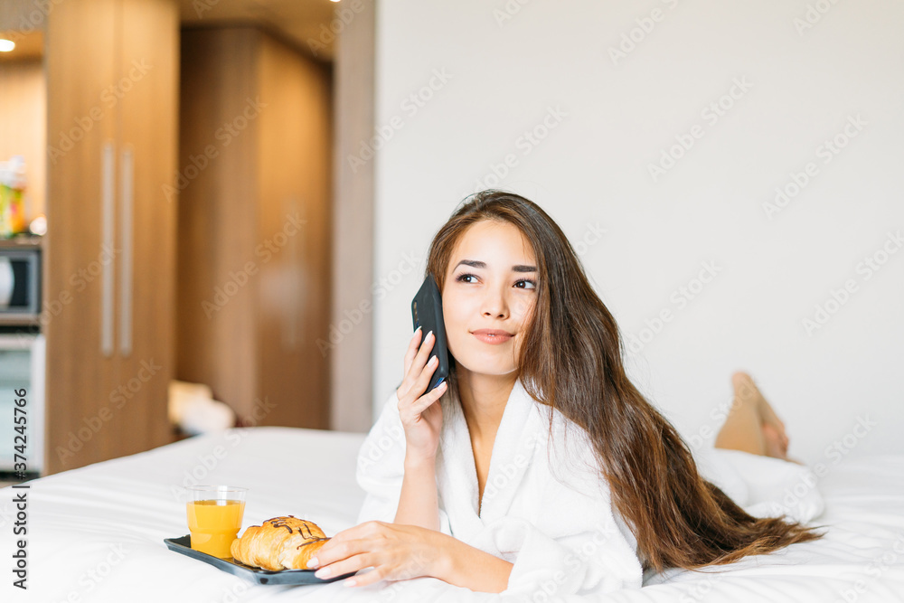 Beautiful young asian woman with long hair in white robe using mobile having breakfast croissant and orange juice in bed of the hotel room