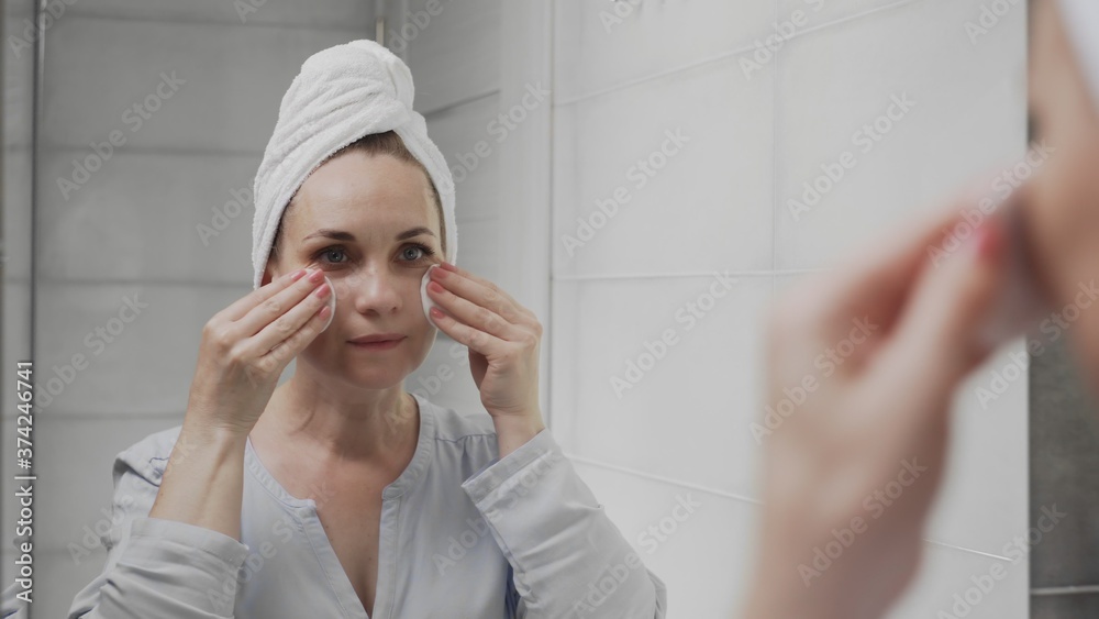 Adult woman with a towel on her head applying lotion and looking in mirror enjoy natural beauty and healthy Adult woman with a towel on her head applying lotion and looking in mirror enjoy natural beauty and healthy